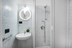 Une salle de bain avec un lavabo blanc, un miroir rond et une douche équipée d’un pommeau de douche à main.