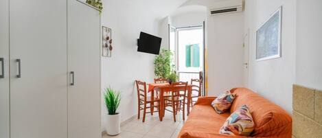 Un espacio habitable compacto con un sofá, una mesa de comedor con sillas, una planta en maceta y un televisor montado en la pared.