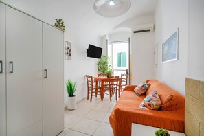 Un espace de vie compact comprenant un canapé, une table à manger avec des chaises, une plante en pot et un téléviseur fixé au mur.