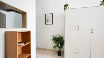 Um quarto com estante de madeira, guarda-roupa branco, planta em vaso e um desenho arquitetônico emoldurado na parede.