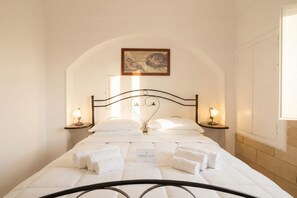 Un lit bien fait, avec du linge de lit blanc et une tête de lit en métal, deux lampes de chevet et une œuvre d’art encadrée au-dessus du lit.