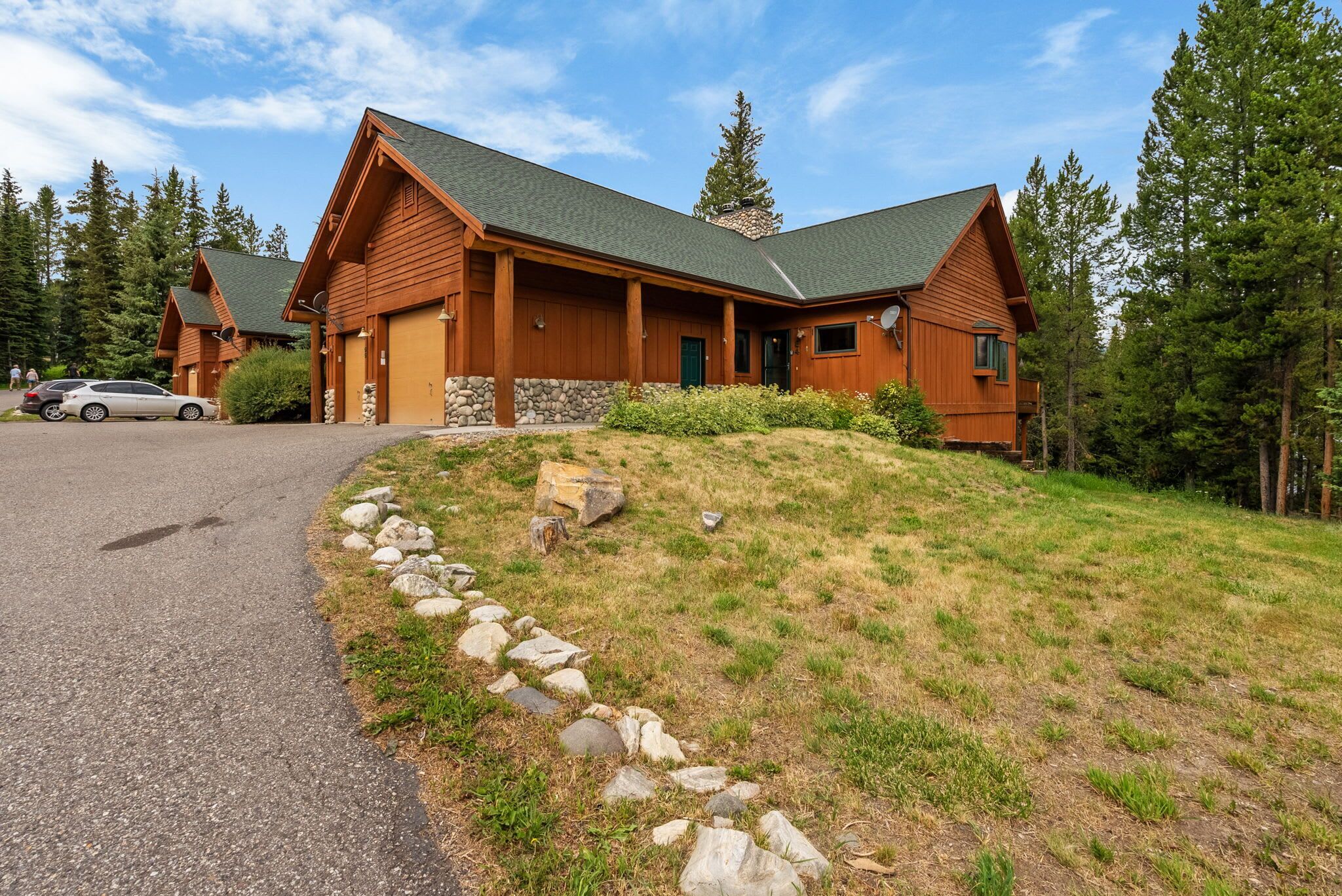 A lodge with a stone foundation, wooden exterior, and a green roof, surrounded by a grassy area and trees.