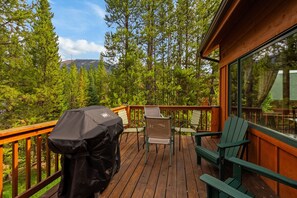 A wooden deck with a grill, chairs, and a view of a forest and mountains.