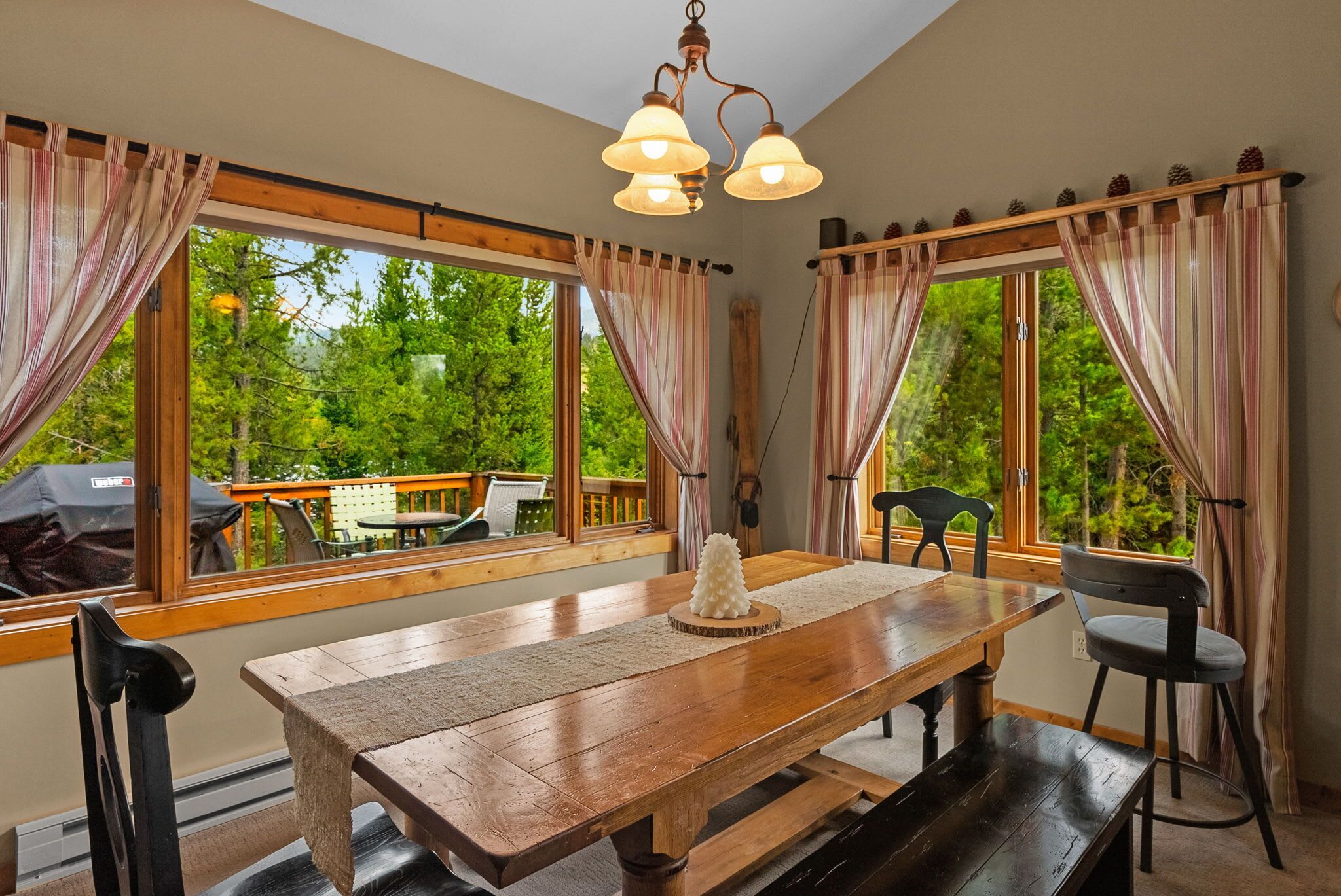 Comedor con mesa de madera, sillas y lámpara de araña. Amplias ventanas con cortinas ofrecen vistas al exterior, con vegetación.