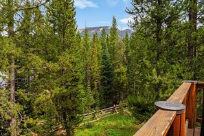 Una terraza de madera con una mesa circular, rodeada de árboles verdes y una montaña al fondo.