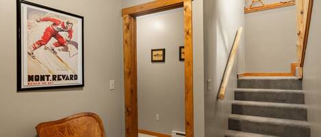 Un pasillo con una puerta de madera, un cartel enmarco, una silla y una escalera.