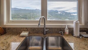 Une cuisine équipée d’un évier en acier inoxydable à deux bacs, d’une fenêtre offrant une vue sur les montagnes, et d’un plan de travail muni d’un distributeur de savon et d’un porte-serviettes en papier.