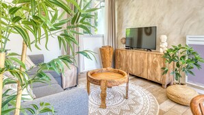 A modern living room with a sofa, a wooden coffee table, a TV on a wooden cabinet, and a large window with curtains. - Villa Indonésie - Domaine Le Sherwood (Notre-Dame-de-Monts)