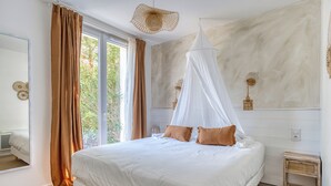 A bedroom with a large bed, a canopy, a bedside table, a mirror, and a view of greenery outside. - Villa Indonésie - Domaine Le Sherwood (Notre-Dame-de-Monts)