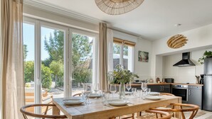 A dining area with a wooden table and chairs, a kitchen in the background, and large windows with a view of trees. - Villa Indonésie - Domaine Le Sherwood (Notre-Dame-de-Monts)