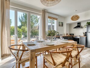 Uma sala de jantar com mesa e cadeiras de madeira, uma cozinha ao fundo, e janelas amplas com vista para árvores.