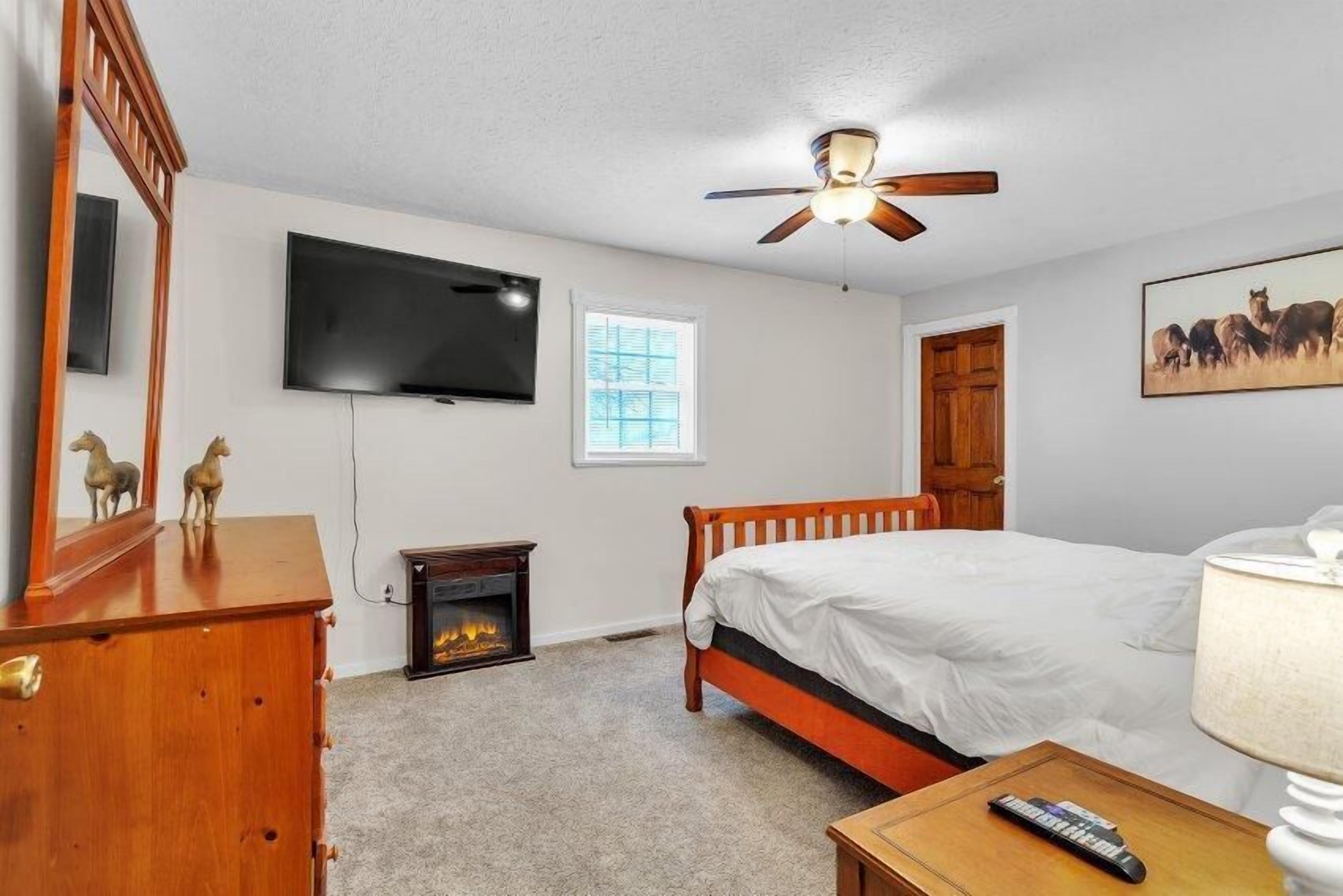 A bedroom with a bed, a television, a ceiling fan, a fireplace, and a wooden dresser.