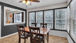 Un coin repas avec une table et des chaises en bois, de grandes fenêtres équipées de stores et un ventilateur de plafond.
