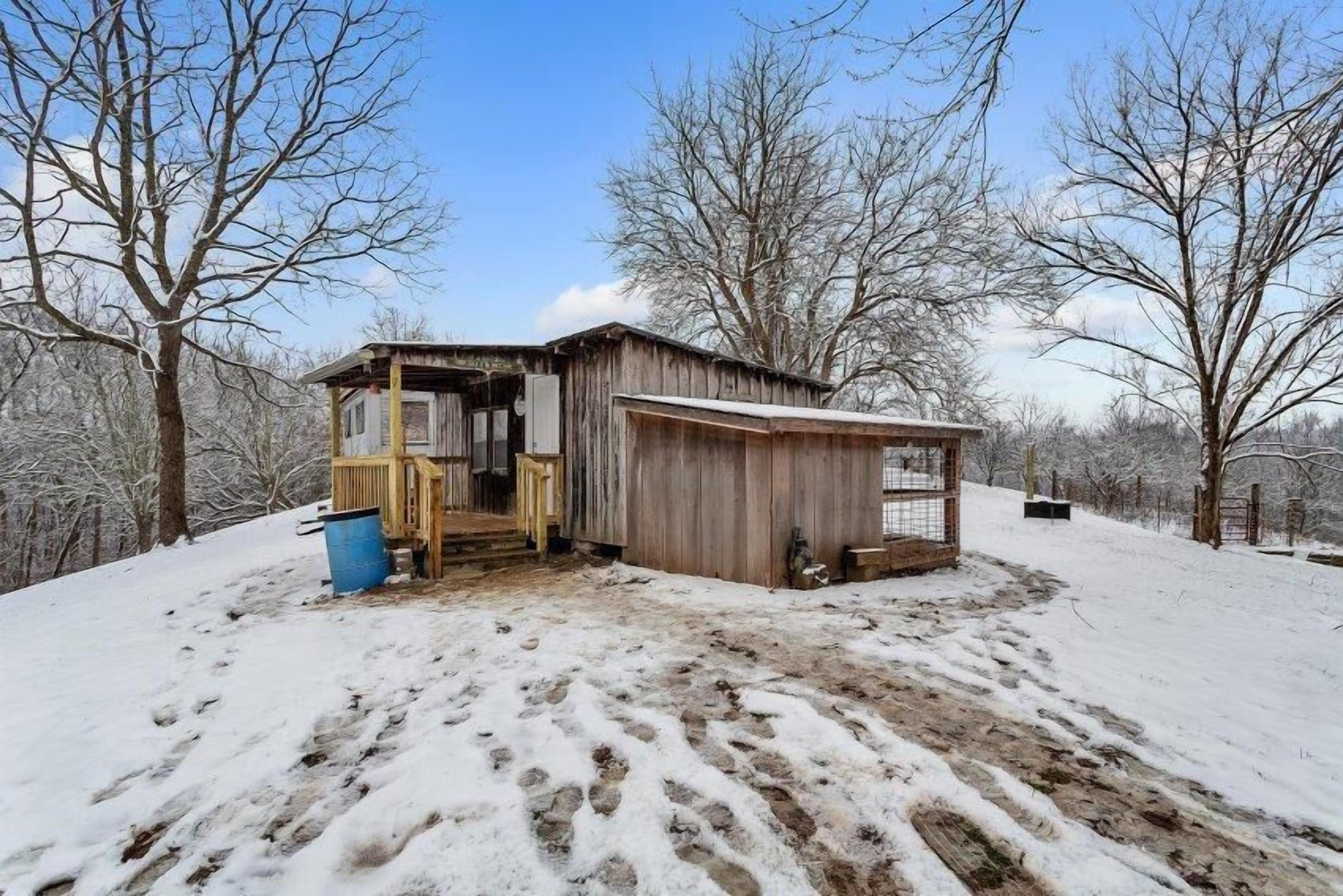 A rustic wooden cabin with a porch and a fenced area, surrounded by snow-covered ground and leafless trees.