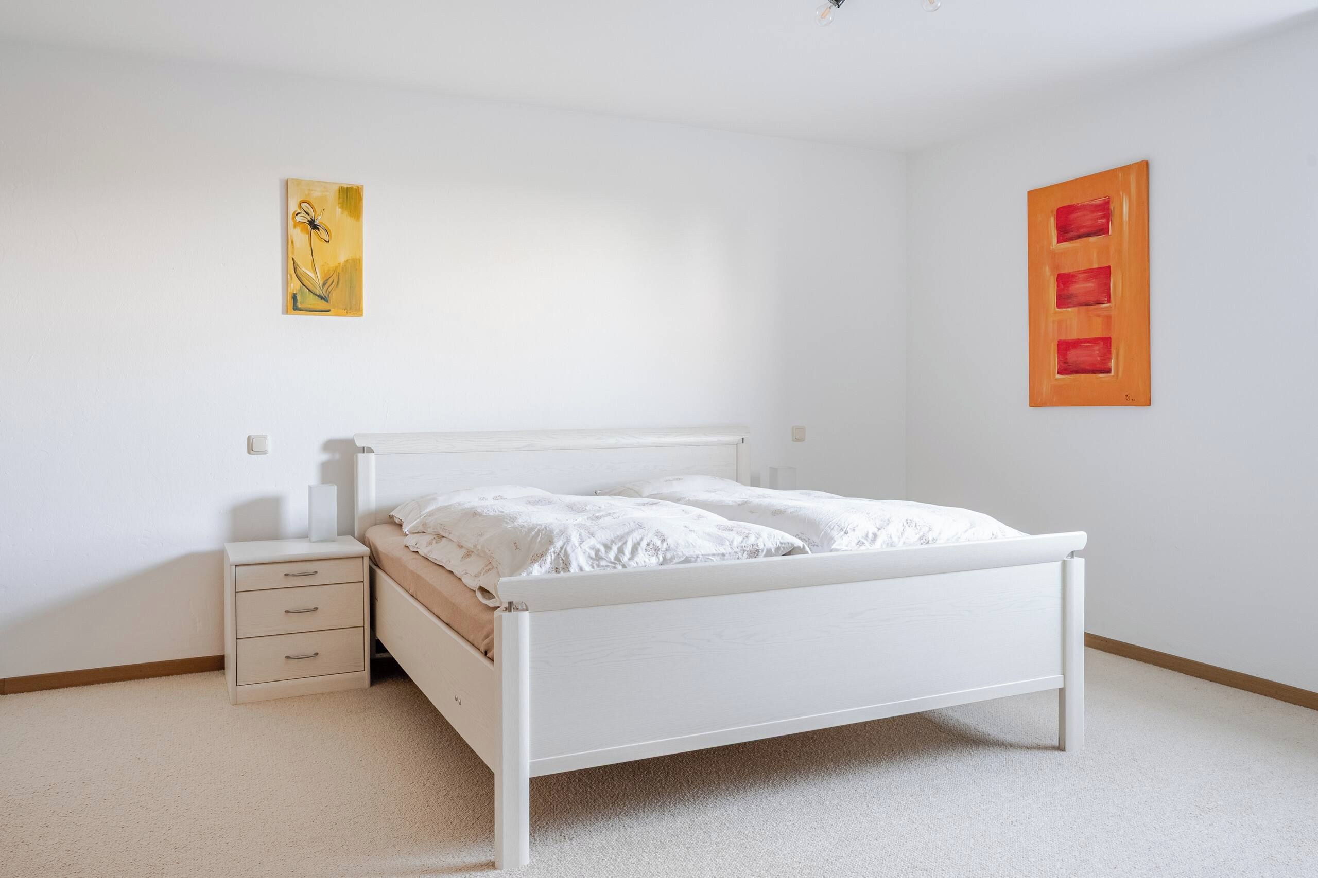 Una camera da letto con un letto bianco, un comodino, un mobiletto e due opere d'arte appese al muro.