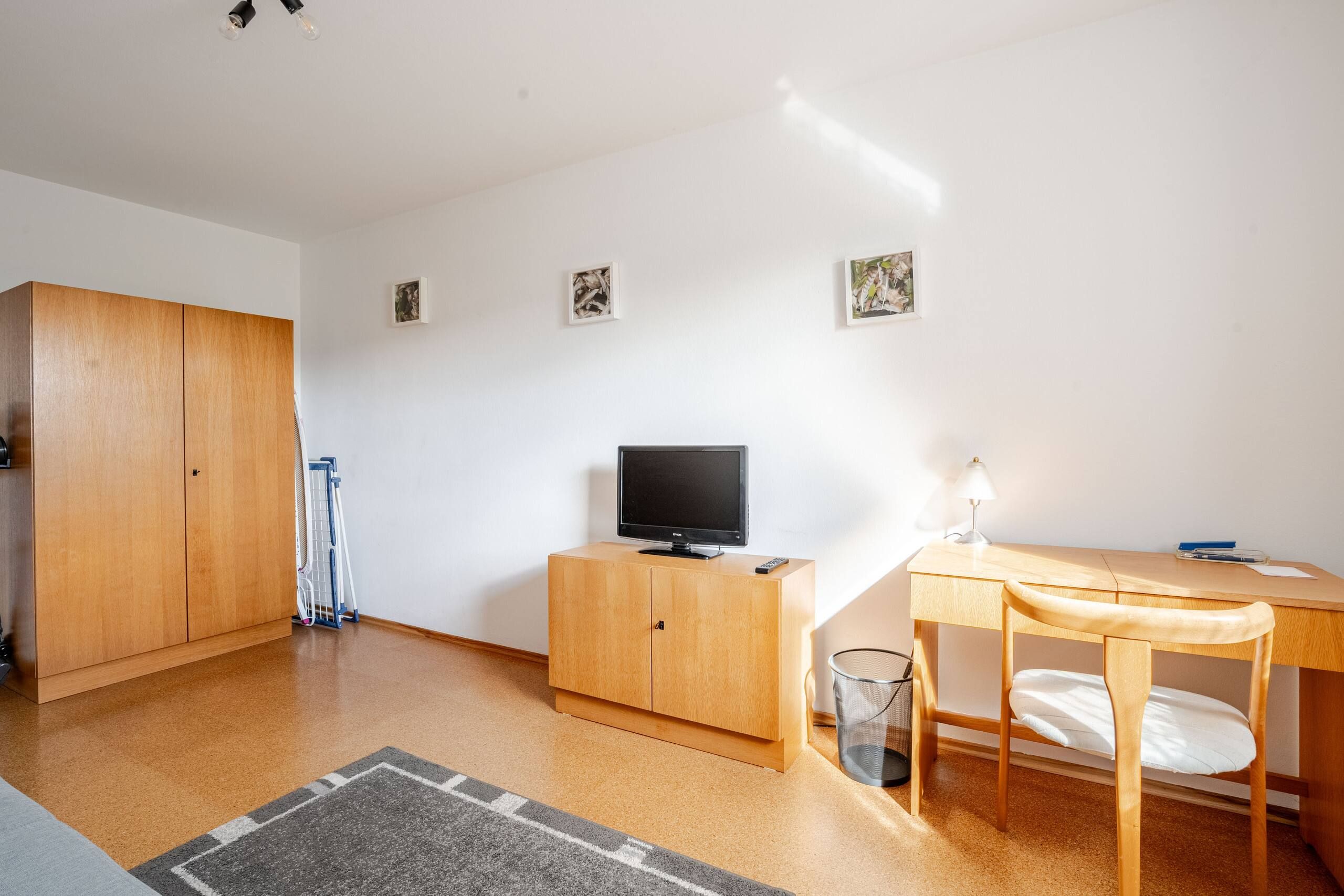 A room with a wooden wardrobe, a television on a wooden cabinet, a desk with a chair, and a small trash bin.