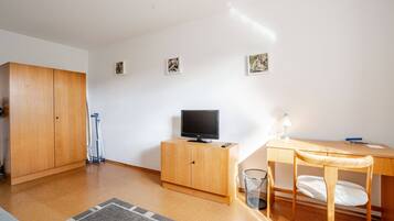 A room with a wooden wardrobe, a television on a wooden cabinet, a desk with a chair, and a small trash bin.