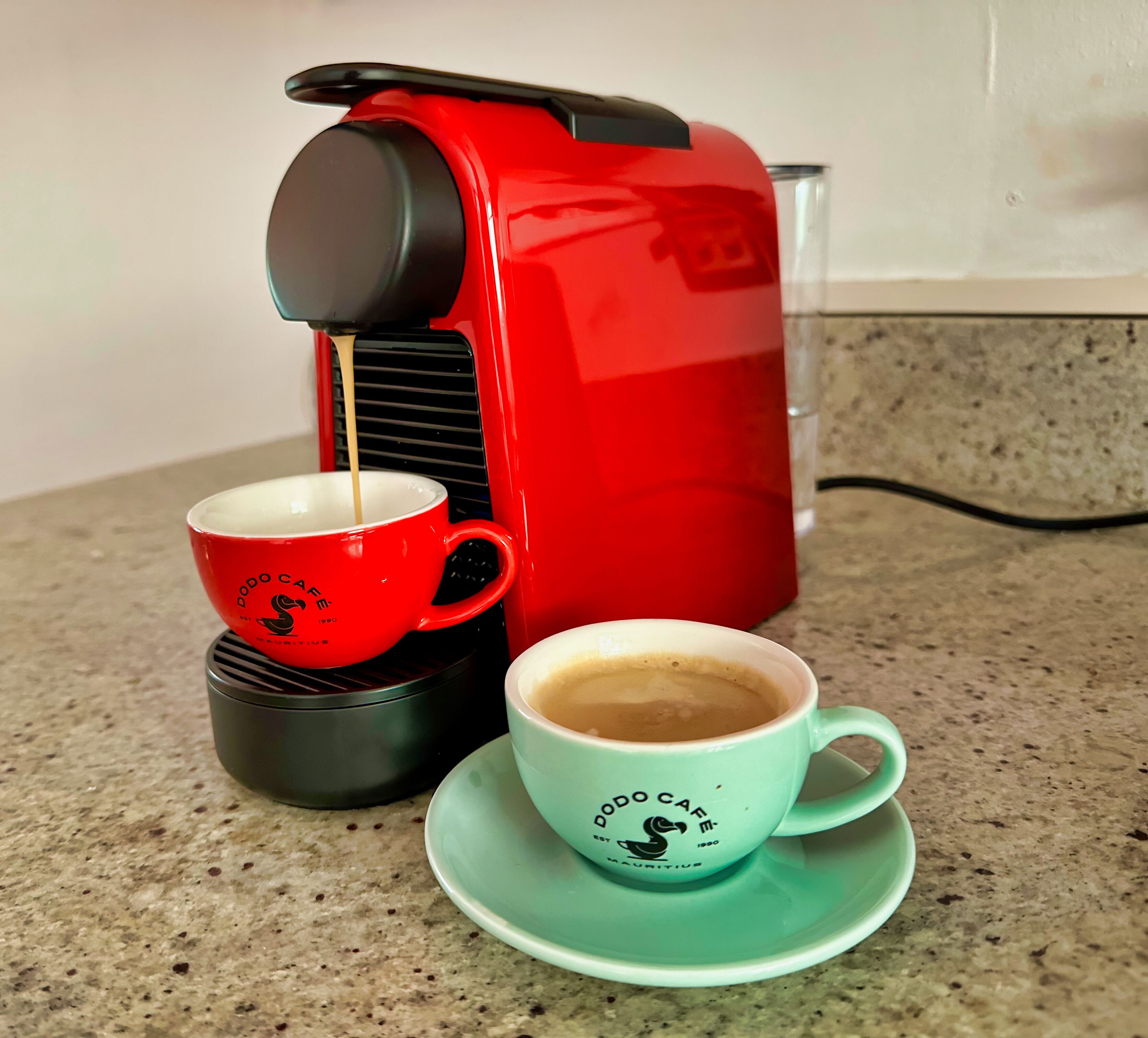 Una caffettiera rossa su un piano di lavoro in cucina, con due tazze, una rossa e una verde, posate sui piattini.