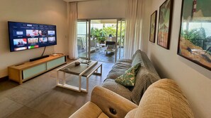 Un salon moderne doté d’un téléviseur à écran plat, d’un ventilateur de plafond, d’un canapé, d’une table basse et d’une porte-fenêtre coulissante donnant sur un espace extérieur avec une terrasse et des plantes.