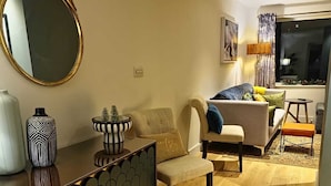 Un salon moderne avec un canapé, un fauteuil et une table basse. Un grand miroir rond est accroché au mur, et une petite table d’appoint est placée à côté, ornée d’objets décoratifs.