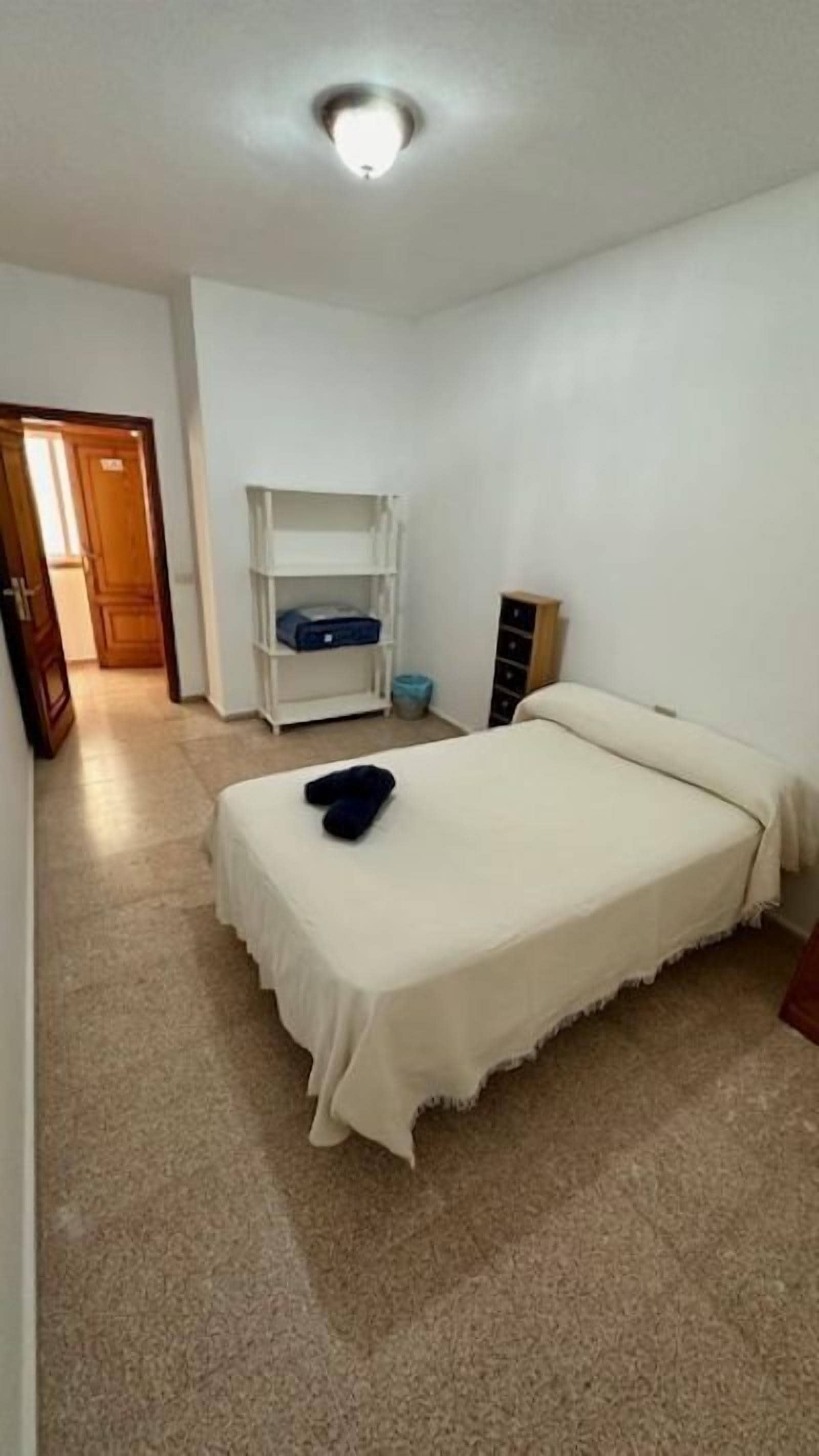 A small, clean room with a single bed, a white shelf unit, and a door leading to another room.