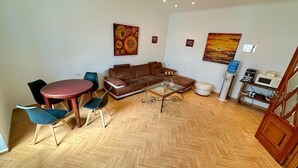 A living room with a brown sofa, a glass coffee table, and a wooden herringbone floor. There are two chairs, a small table, and a microwave on a shelf.