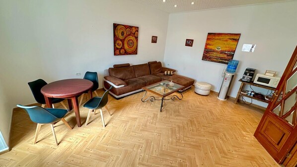 A living room with a brown sofa, a glass coffee table, and a wooden herringbone floor. There are two chairs, a small table, and a microwave on a shelf.