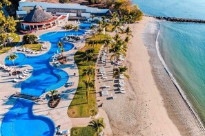 Luchtbeeld van een resort met meerdere zwembaden, een strand en een gebouw met rieten dak.