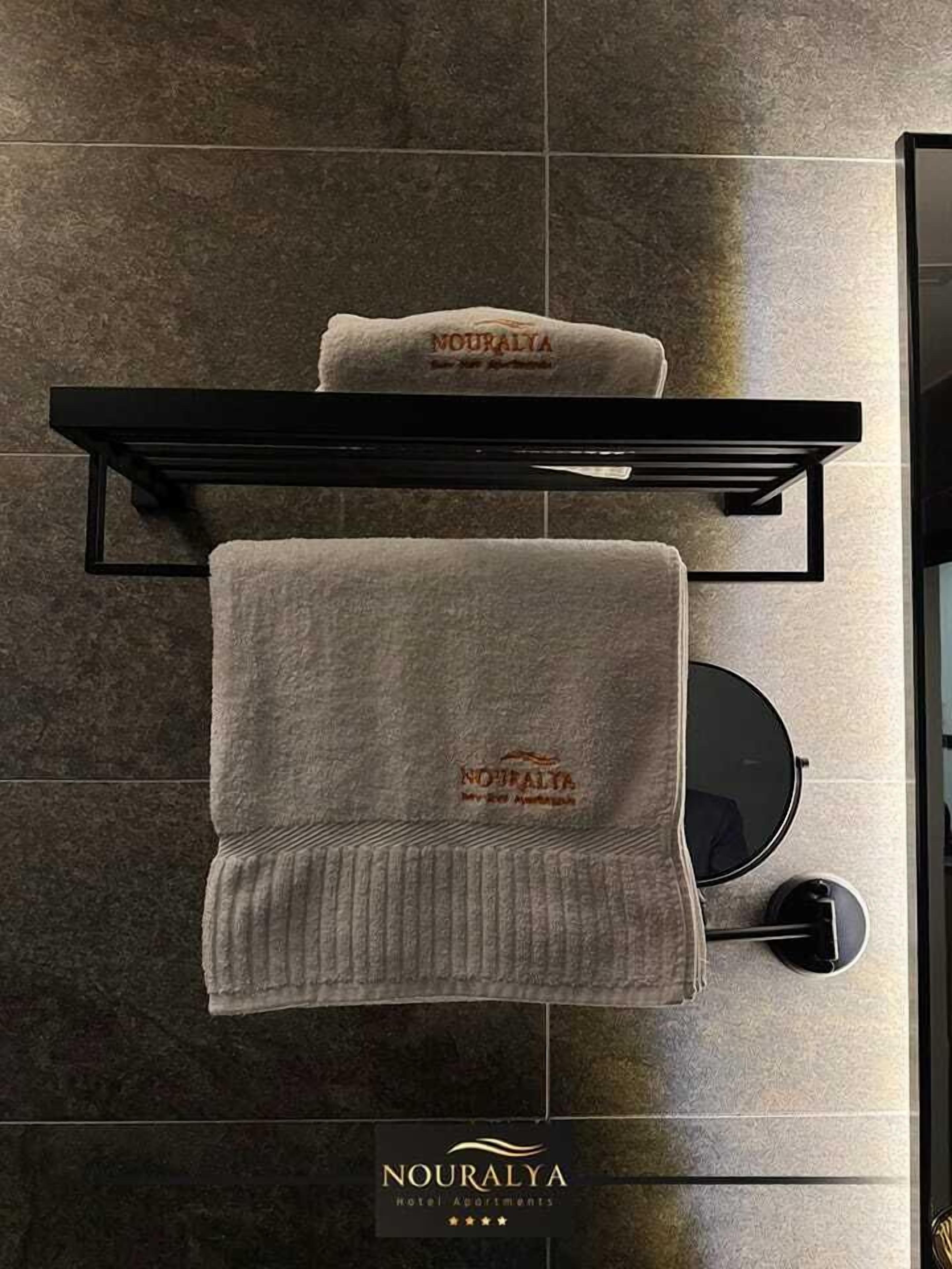 A bathroom with a towel rack holding two towels, one with 'NOURALYA' embroidered on it, and a sign below stating 'NOURALYA Hotel Apartments'.
