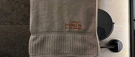 A bathroom with a towel rack holding two towels, one with 'NOURALYA' embroidered on it, and a sign below stating 'NOURALYA Hotel Apartments'.