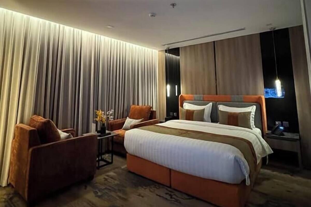 Una habitación de hotel moderna con una cama grande, una zona de estar con una silla y una mesita, y un televisor en la pared.