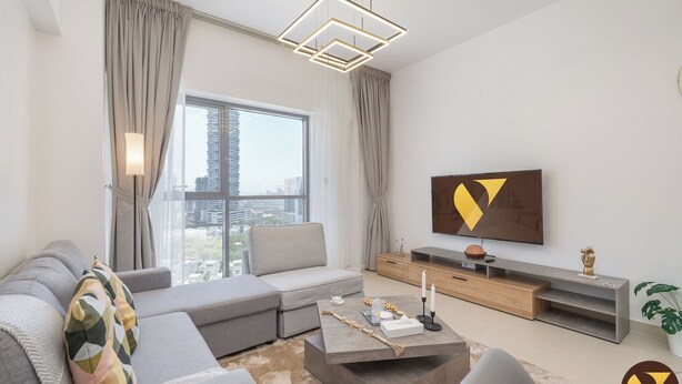 A modern living room with a grey sectional sofa, a coffee table, and a flat-screen TV mounted on the wall. - Brand New with Relaxing Pool & Kids Area (Dubai)