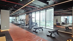 A modern gym with cardio machines, weightlifting equipment, and large windows offering views of the city. - Brand New with Relaxing Pool & Kids Area (Dubai)
