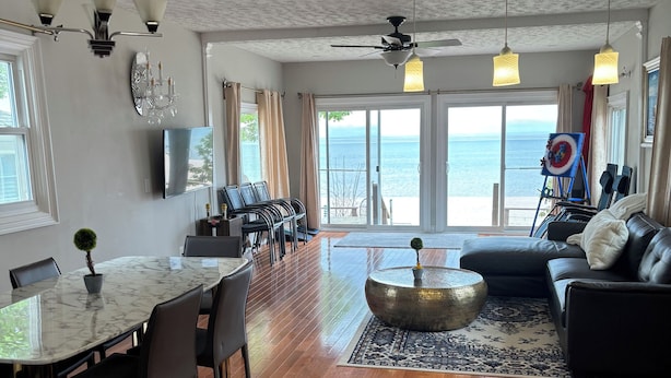 A spacious living area with a wooden floor, a dining table with chairs, a black leather sofa, and a view of the sea through large windows.