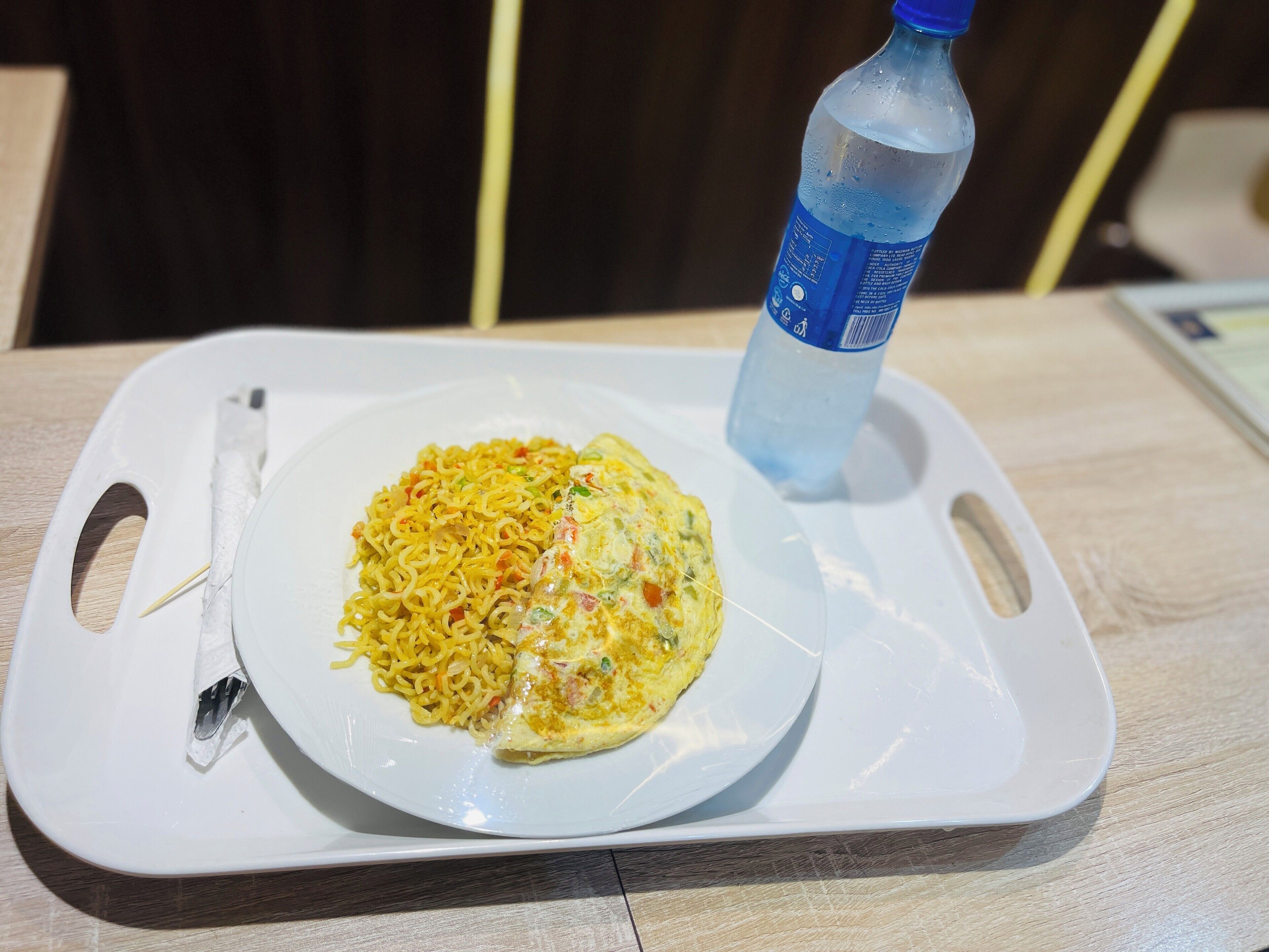 Un vassoio con una porzione di noodles e un'omelette, una forchetta e una bottiglia d'acqua su un tavolo di legno.