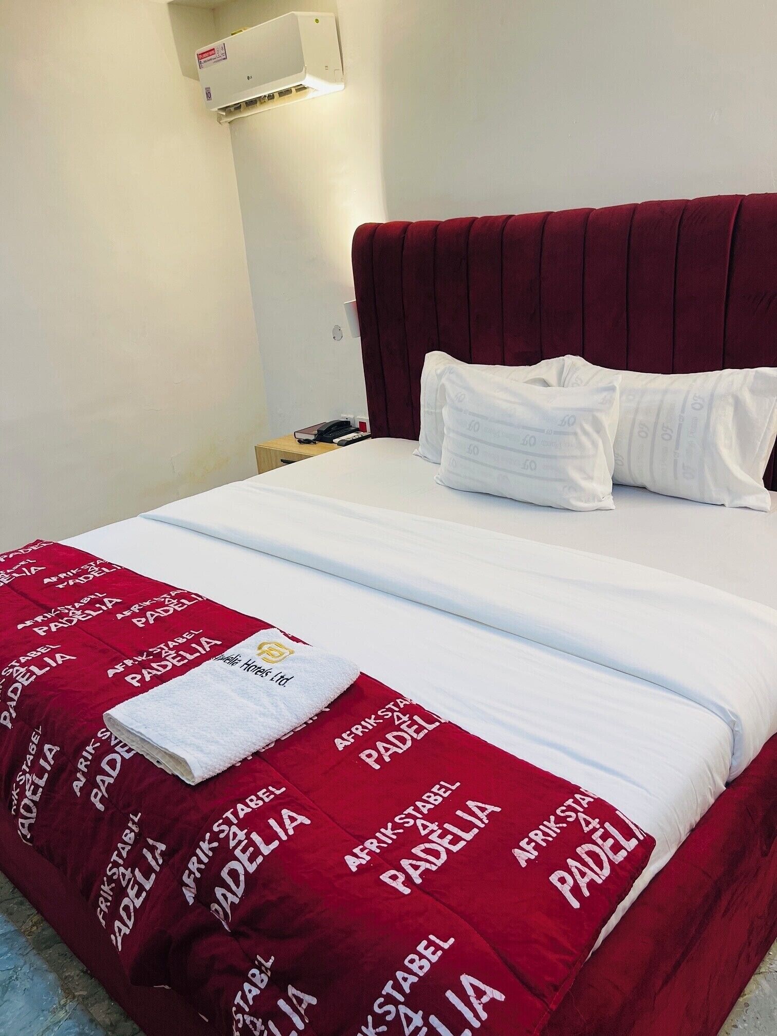 A hotel room with a bed, white linens, a red blanket with 'Afrikstabel Padelia' branding, a white pillow, and a wall-mounted air conditioner.