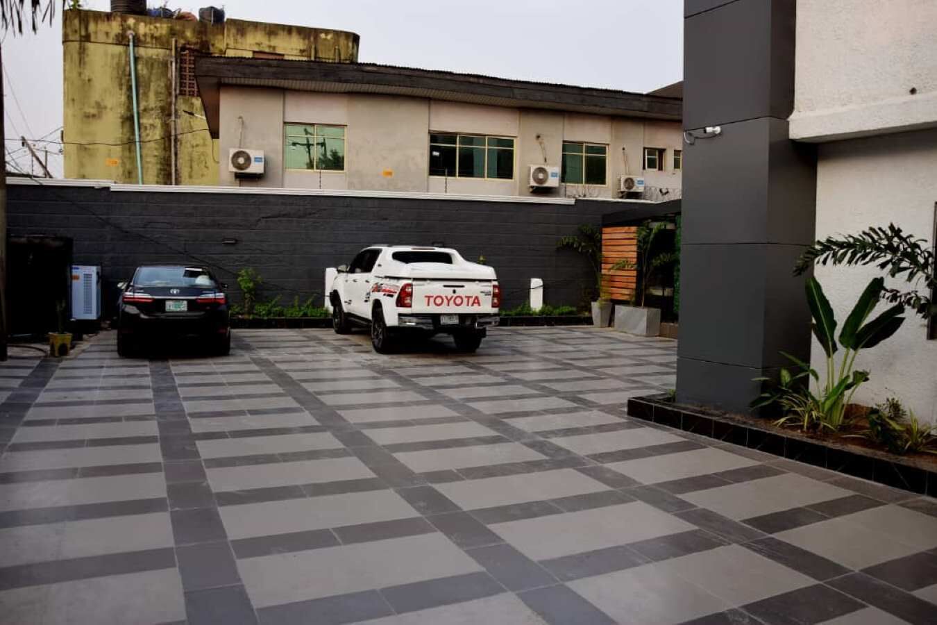 A parking area with two vehicles, a white pickup truck and a black car, parked in front of a building with a modern design and large windows.