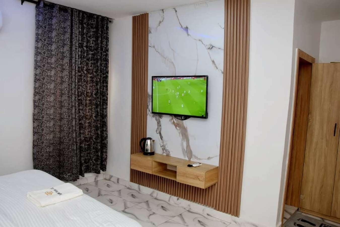 A hotel room with a bed, a television mounted on a marble wall, a wooden shelf with a kettle, and a wardrobe.