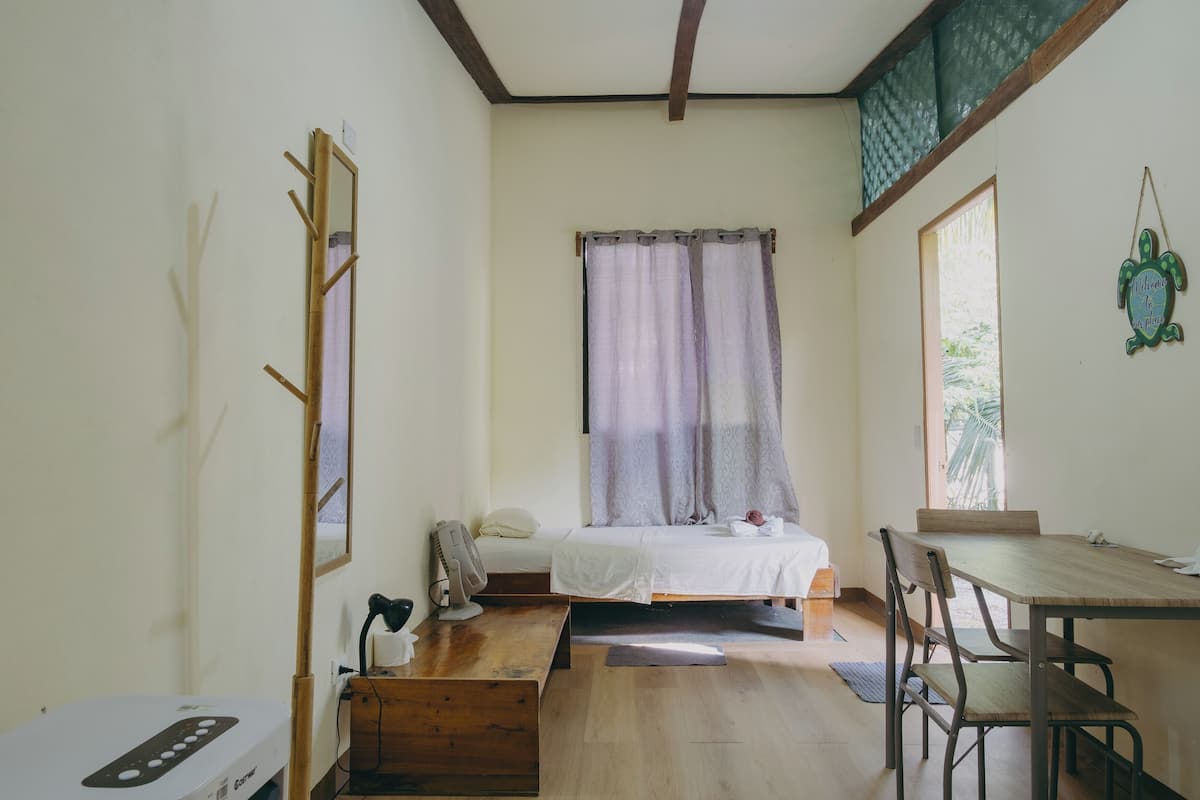 A small, well-lit room with a bed, a desk, and a chair. There is a window with curtains, a wall-mounted mirror, and a small table with a lamp.