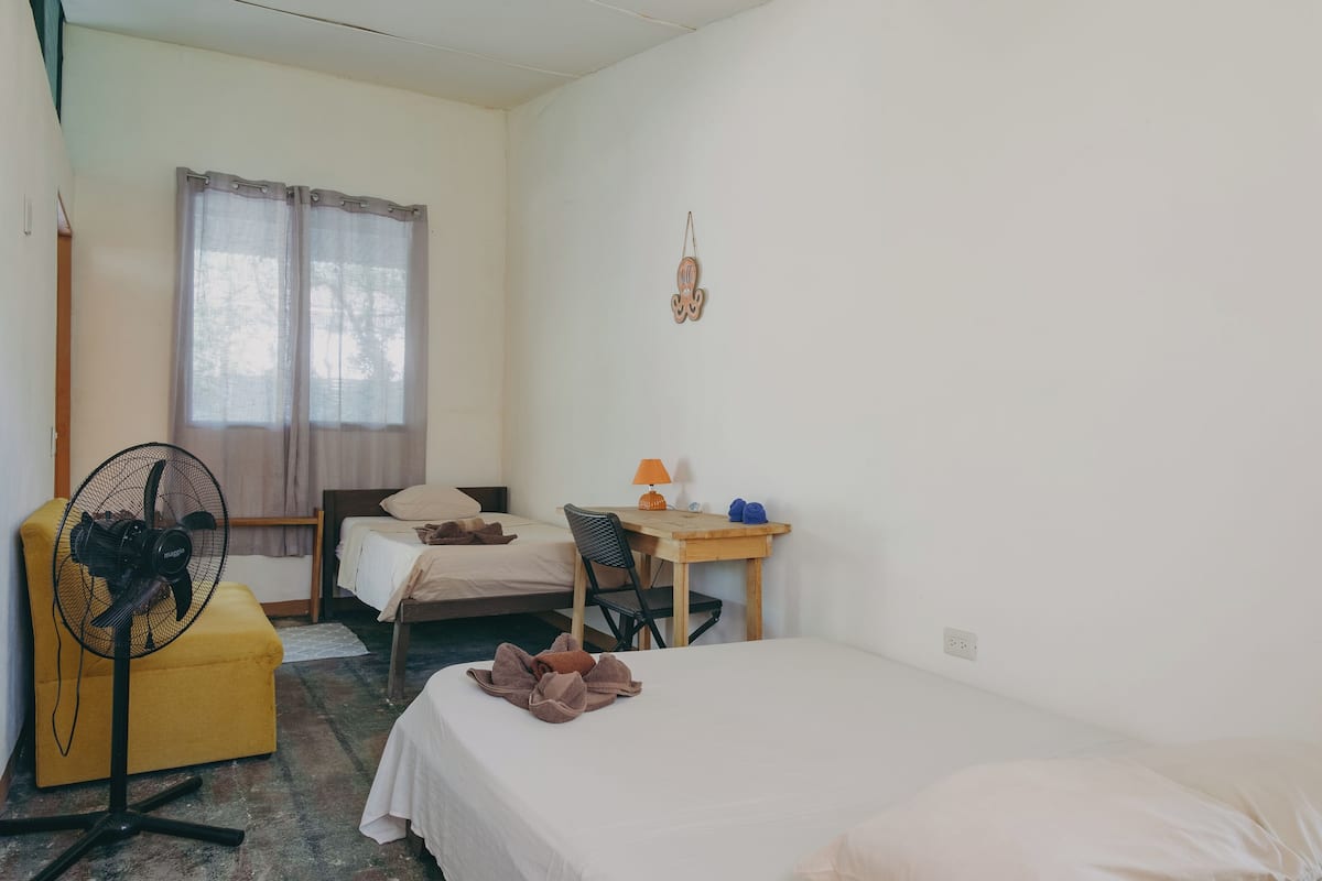 A room with two beds, a desk with a lamp, a chair, a fan, and a window with curtains.