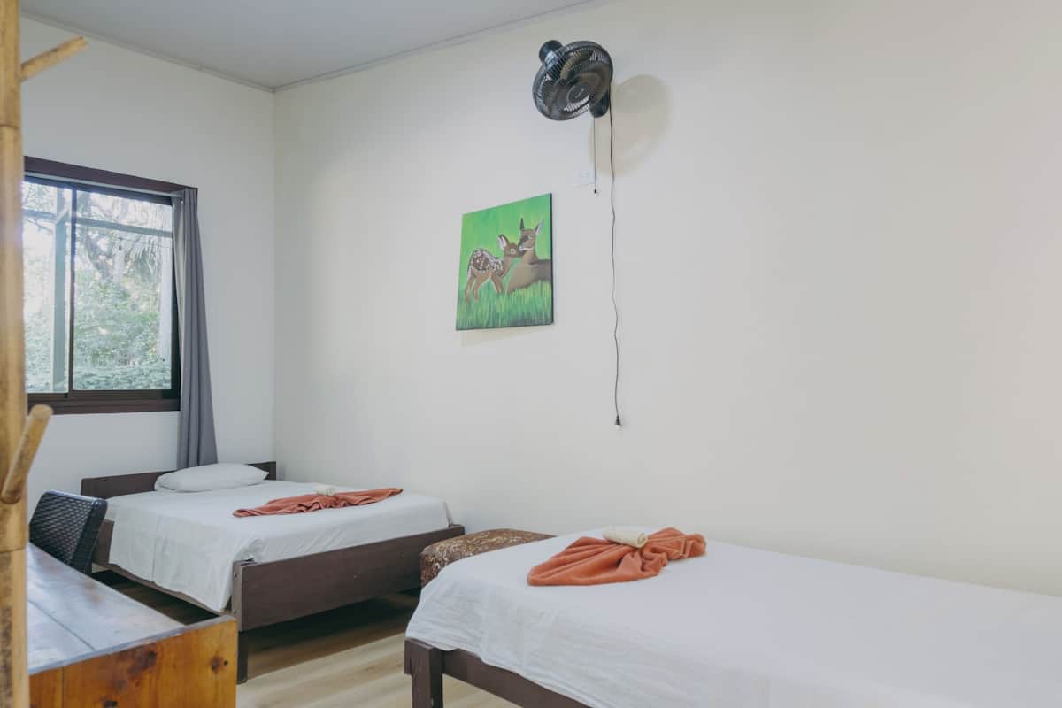 A room with two single beds, a wooden desk, a window with curtains, a wall-mounted fan, and a painting of deer.