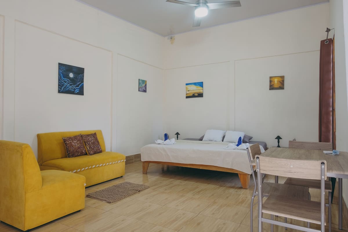 A room with a bed, a yellow sofa, a dining table with chairs, and artwork on the walls.