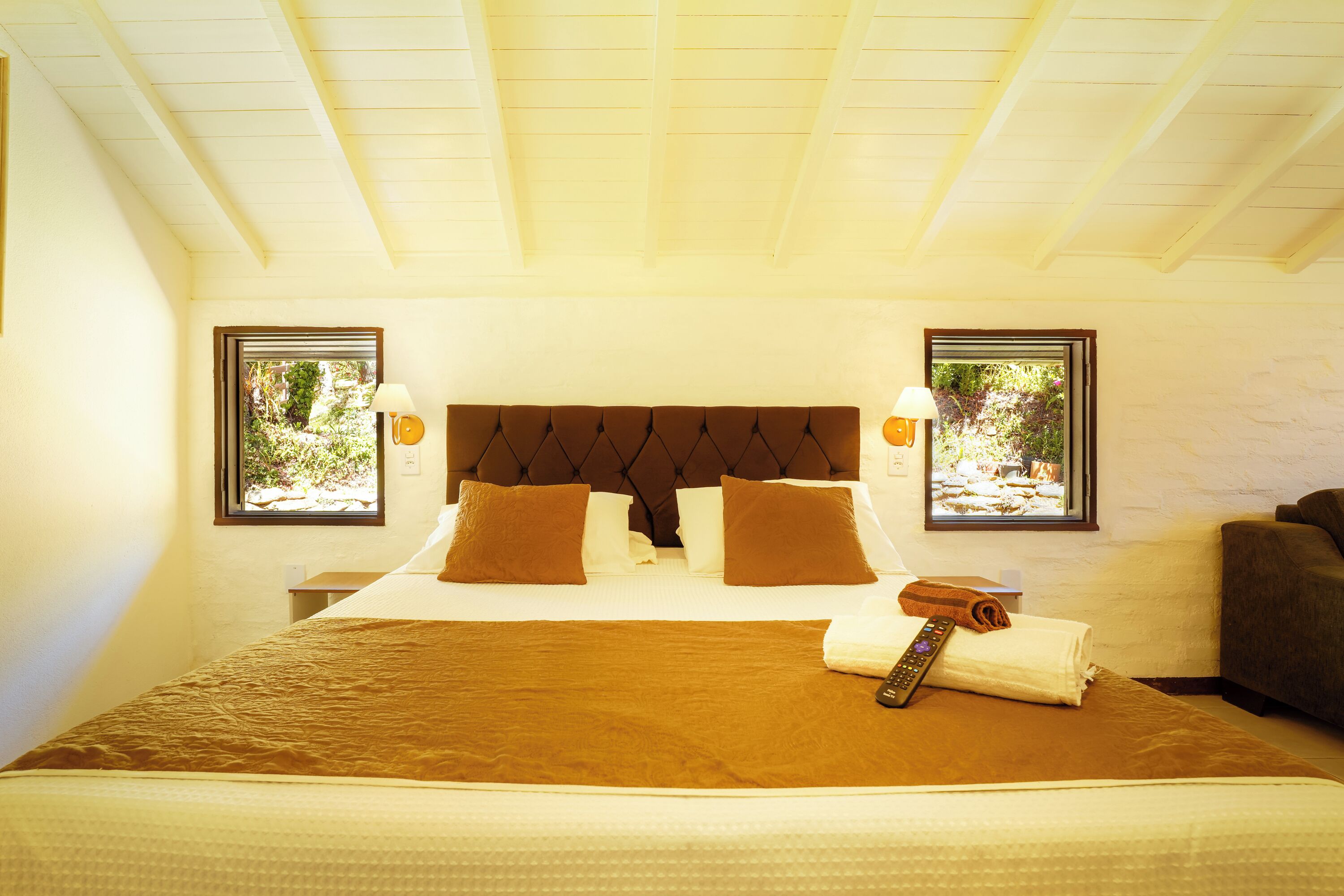 A hotel room with a large bed, two bedside lamps, a headboard, and a view of greenery through windows.