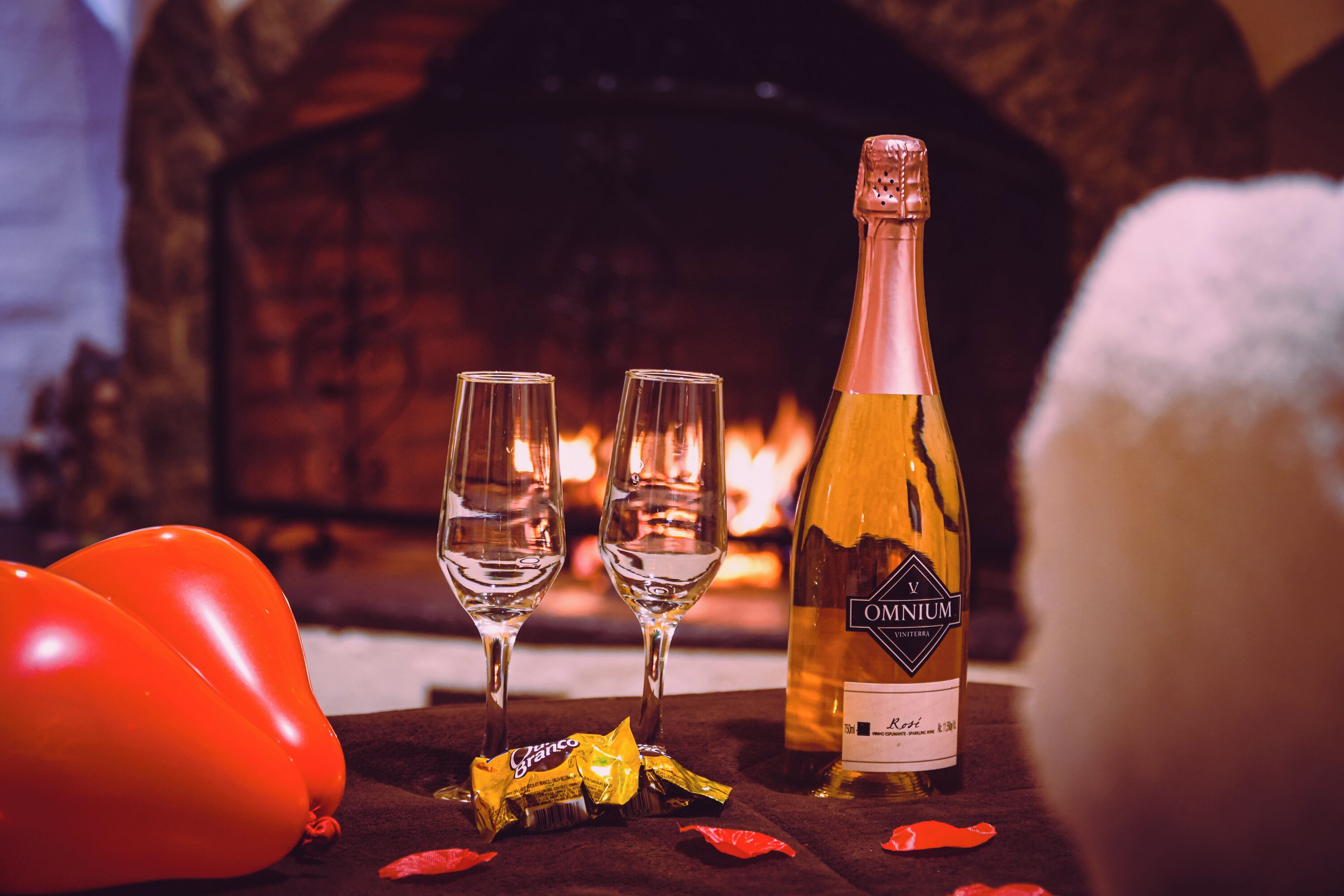 A bottle of wine, two glasses, and red balloons on a table with a fireplace in the background.