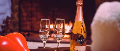 A bottle of wine, two glasses, and red balloons on a table with a fireplace in the background.