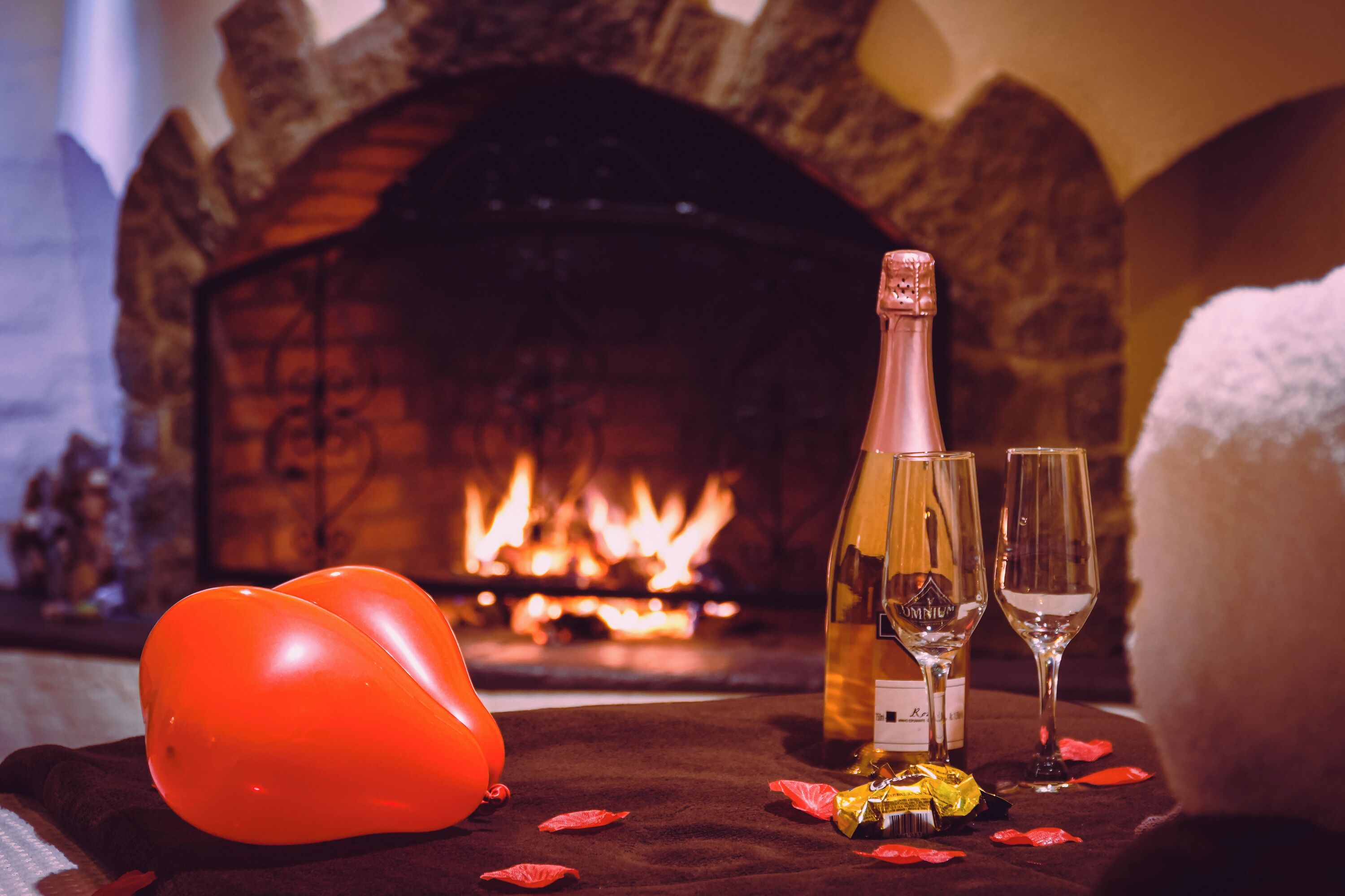 A fireplace with a lit fire, a bottle of wine, two wine glasses, and a heart-shaped balloon on a table.