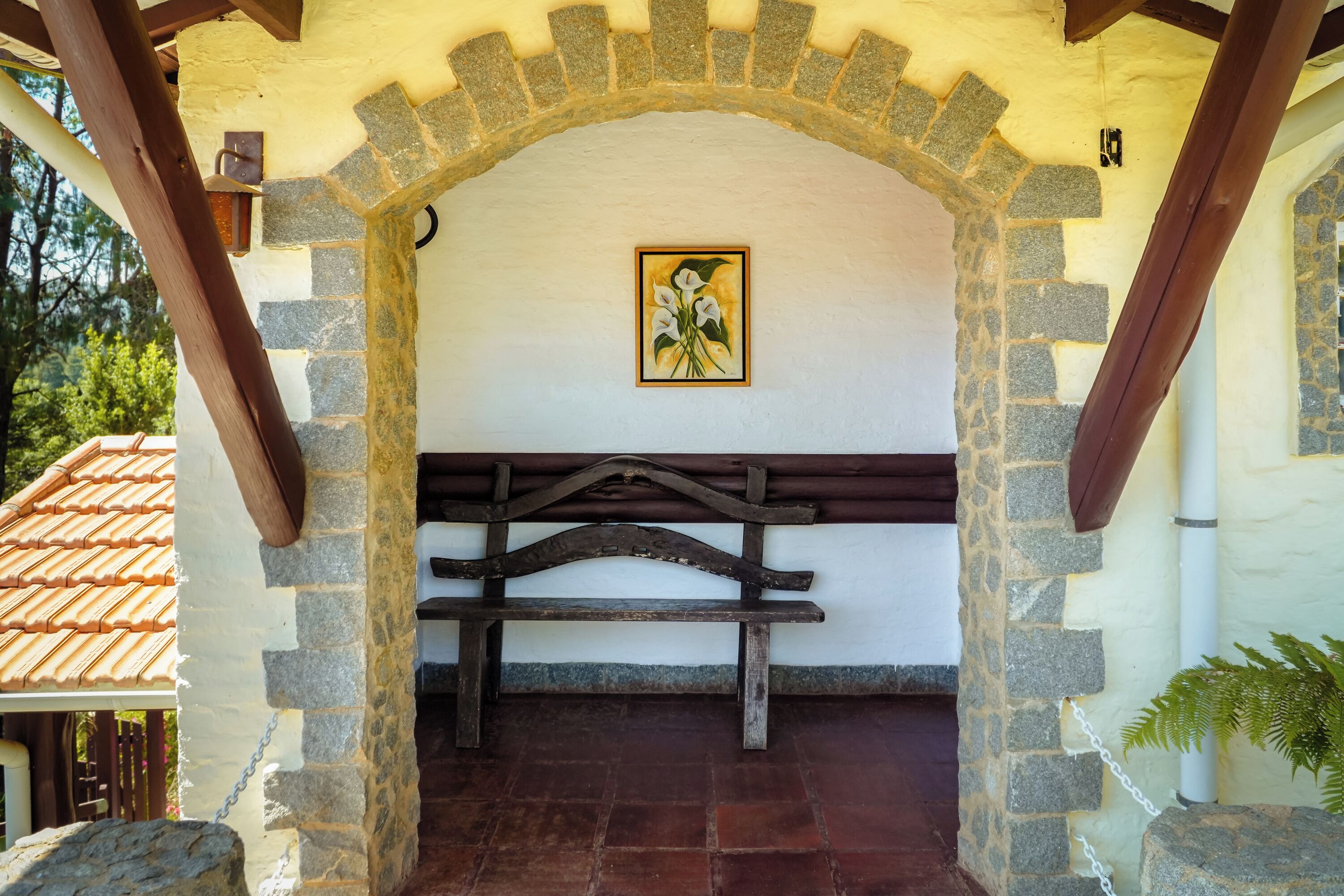 An arched stone structure with a wooden bench, a framed picture on the wall, and a stone wall with a stone bench on the right.