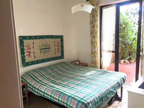 Relax in a cozy room with a single bed, featuring a checkered bedspread and a wall-mounted artwork above. Enjoy the sliding glass door leading to an outdoor area with lush greenery.