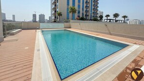 A rooftop swimming pool with a wooden deck, surrounded by high-rise buildings and palm trees. - Jeewar Brand New Relaxing Pool Kids Area (Dubai)