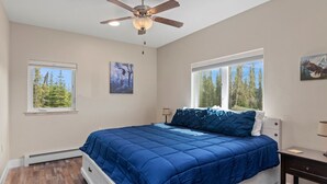 A bedroom with a large bed, two windows with forest views, a ceiling fan, and artwork on the walls. - Modern Alaskan Retreat (Soldotna)